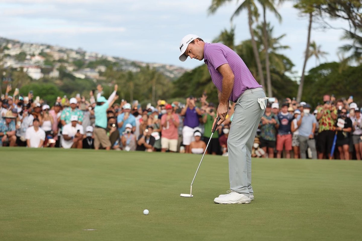 A golfer wearing a pink shirt, white hat and gray pants gets ready to hit the golf ball while people cheer in the background.