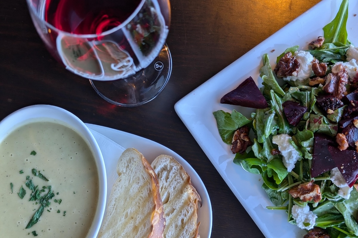 A mixed green salad on a square white plate, a bowl of a light yellow soup with herb garnish and bread on the side and a glass of red wine sit on a table.