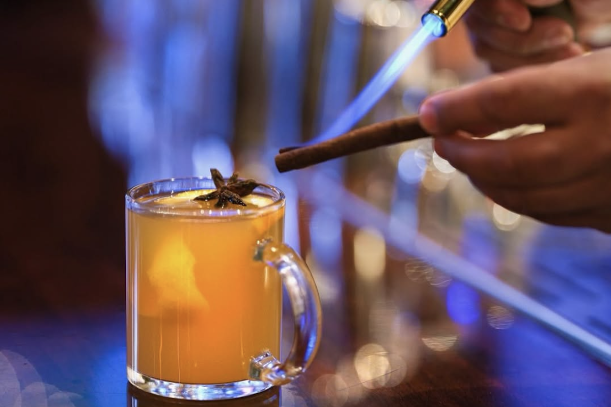 A hot toddy in a glass mug sits on a bar as someone uses a blowtorch on a cinnamon stick above the drink.