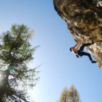 A man dangles high up on the side of a rocky edge, as he rock climbs against a clear blue sky.