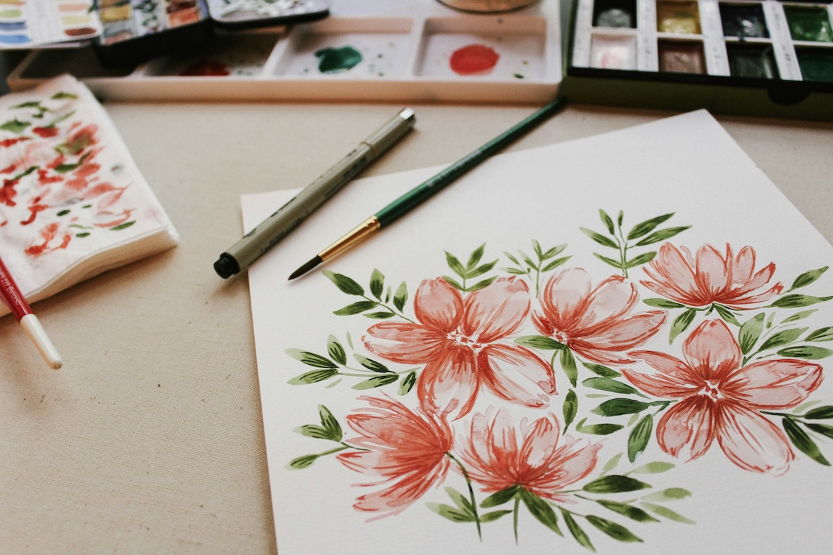 A piece of paper with watercolored pink flowers and green leaves, with the paint palette and some brushes in the back.