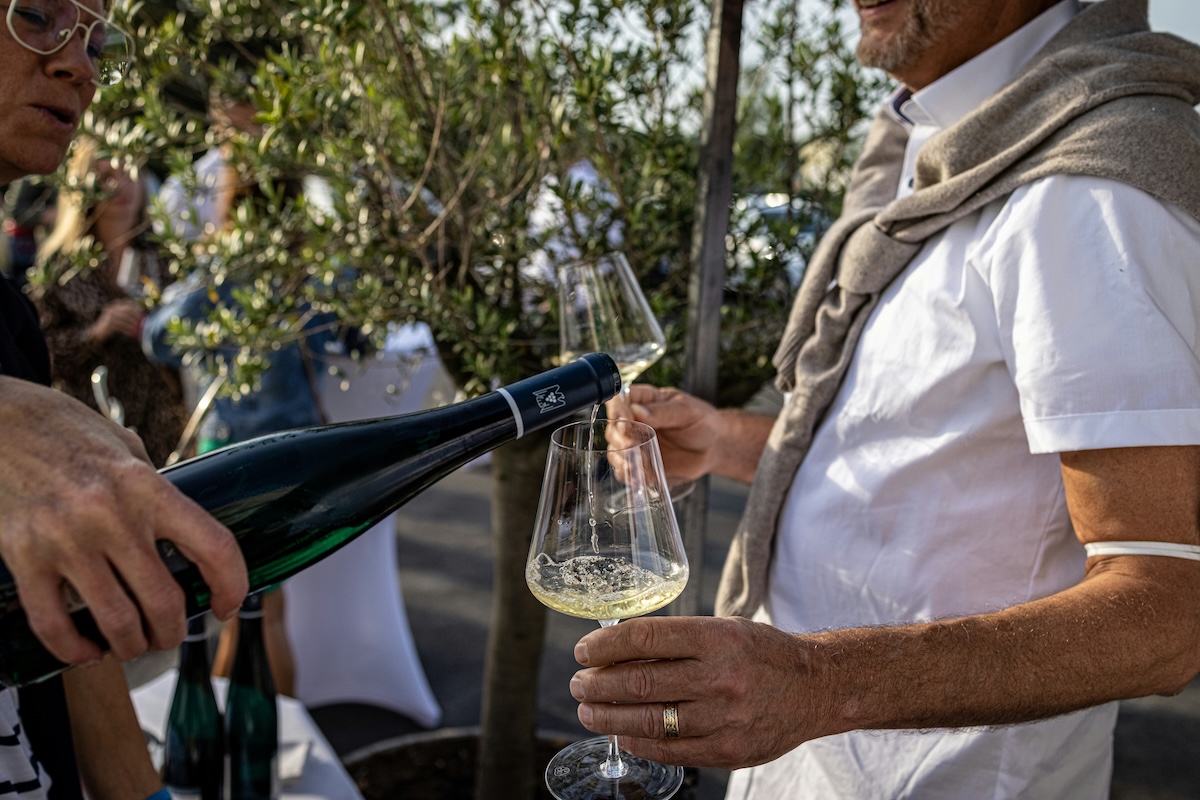 Outside, a man pours wine from a dark bottle into a wine glass held by a man wearing a white T-shirt and gray sweater tied around his neck.