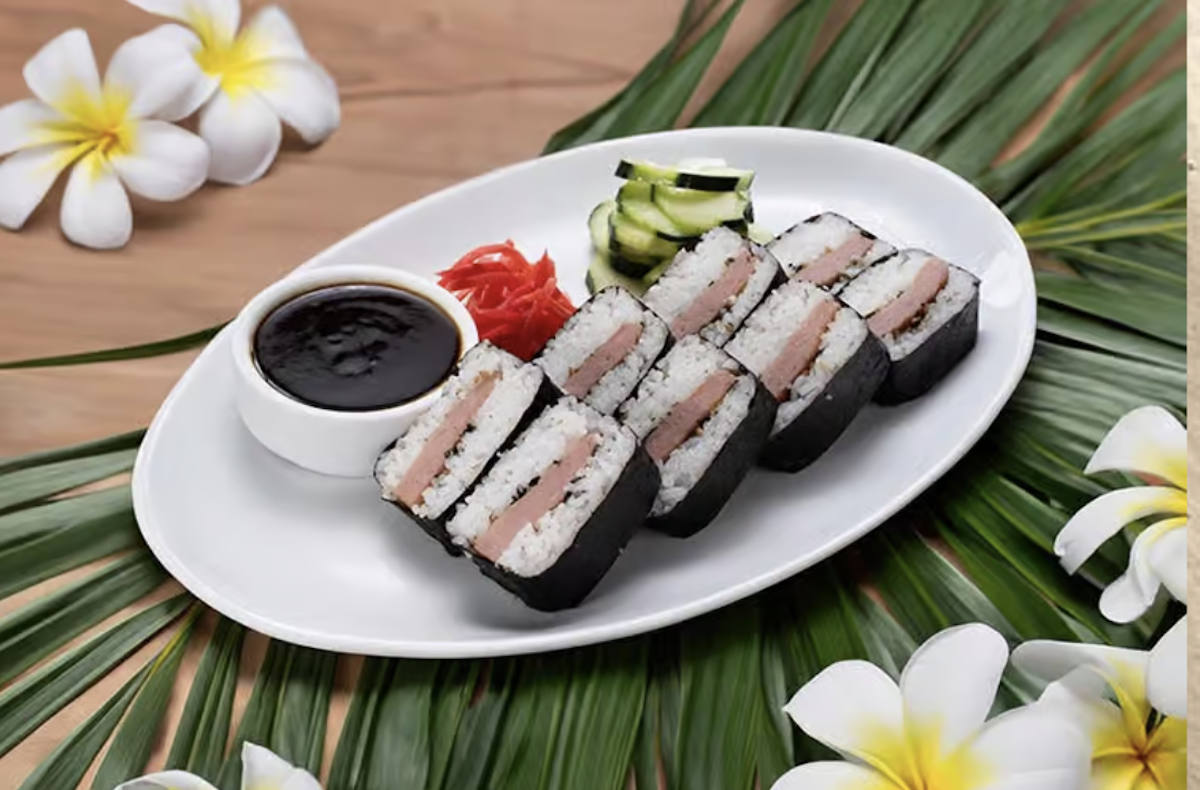 A plate of slices of SPAM musubi with dipping sauce and sides of pickled onions and cucumber, all on a bed of palm leaves decorated with plumeria flowers.