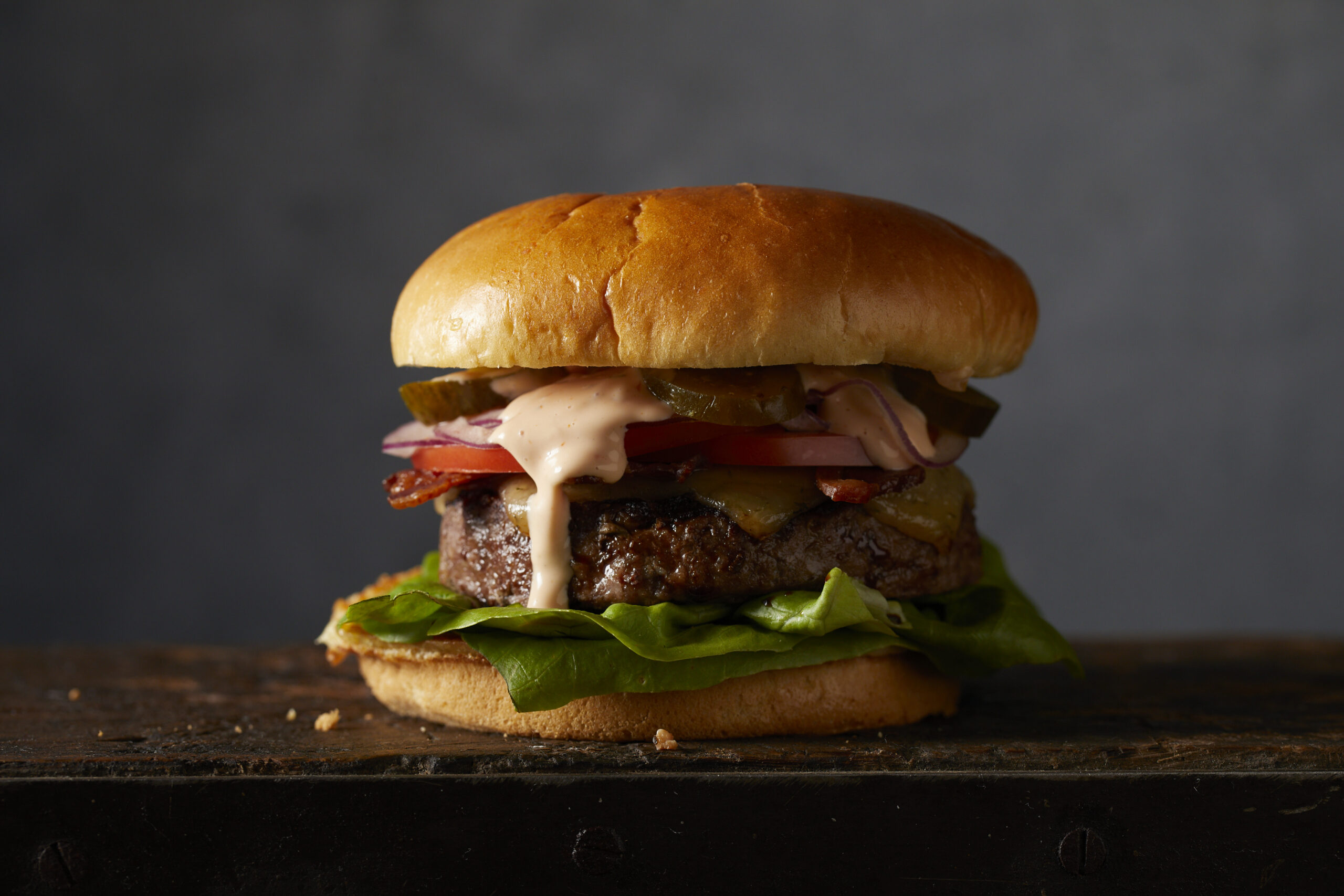 A burger sitting on a table with lettuce, tomato, pickles, red onion and sauce dripping out and down the side.