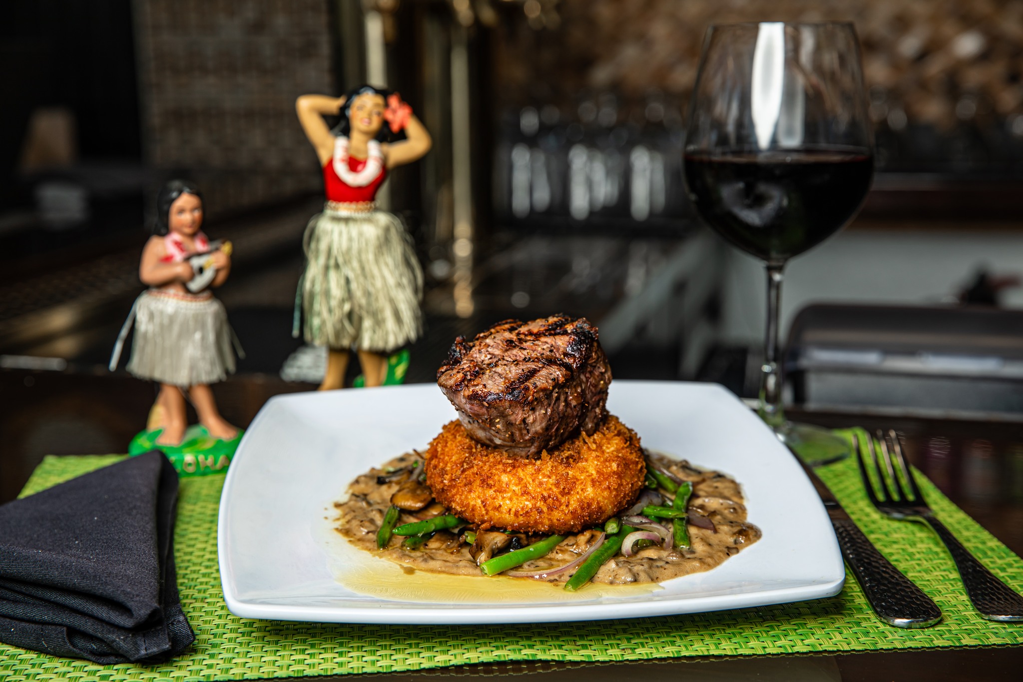 A plate with sauce, mushrooms, onions, asparagus, a breaded piece of something and a steak on top. A couple of hula girl statues are in the background.