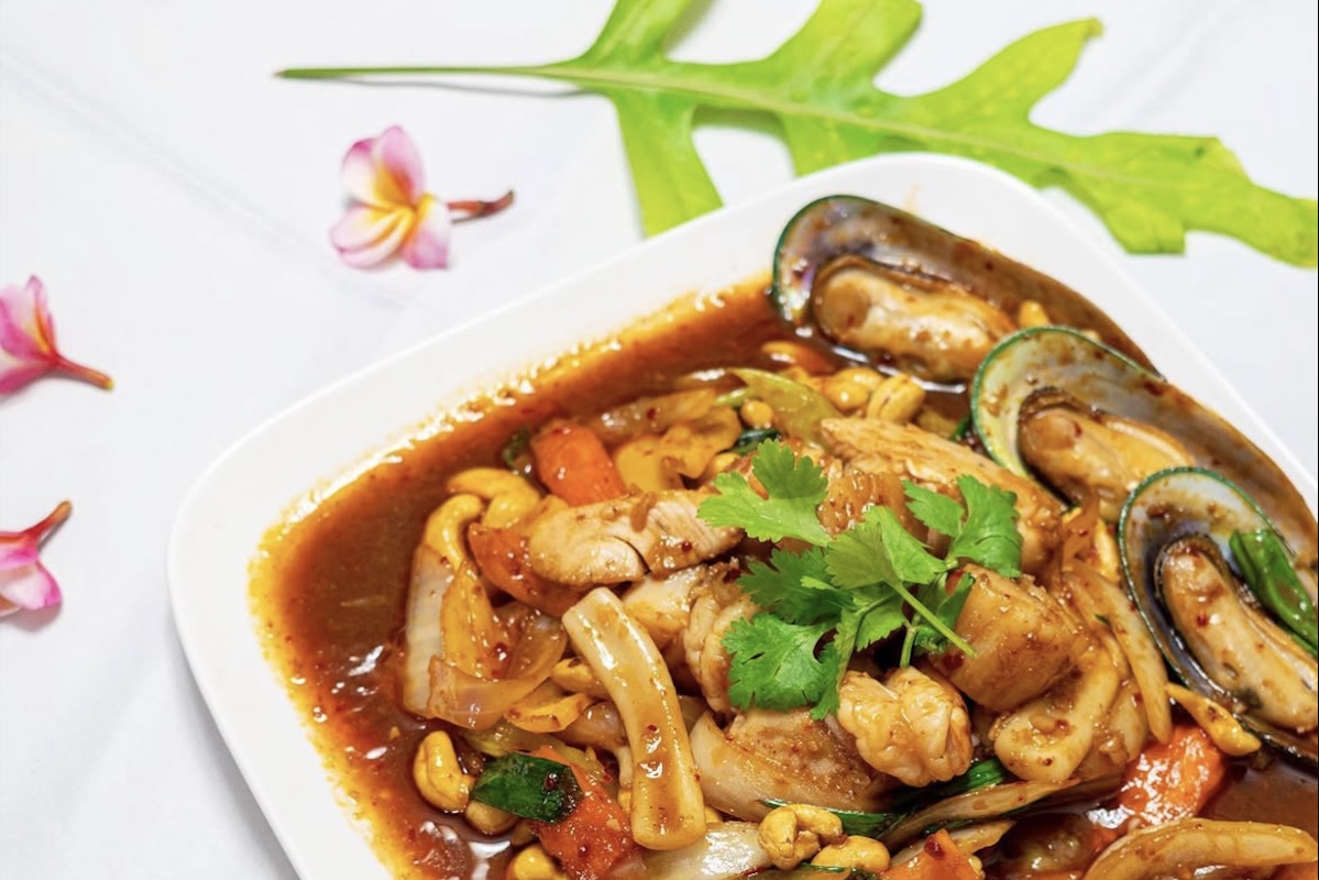 A plate with meat, veggies and seafood doused in a rich reddish brown sauce sit on a table decorated with pink plumerias and a green leaf.