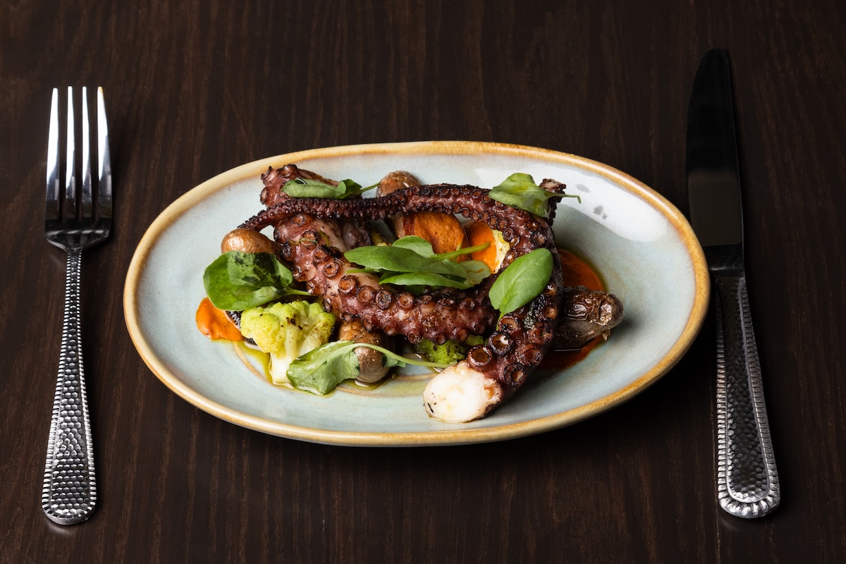 A white plate with roasted octopus accompanied by spinach, cauliflower and roasted potatoes.