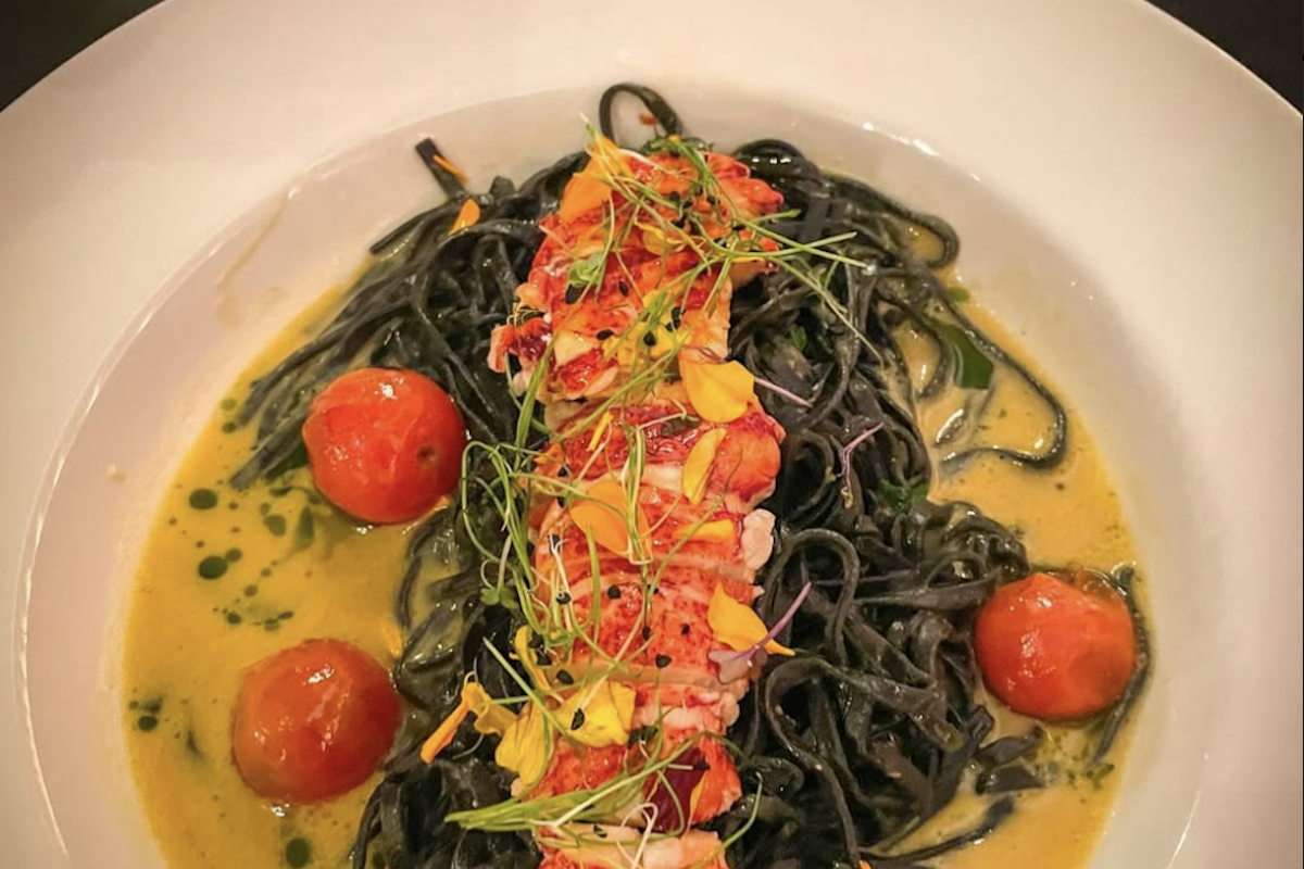 A plate with a bed of black pasta in yellow sauce and topped with sliced seafood, flower petals, sprouts and tomatoes.