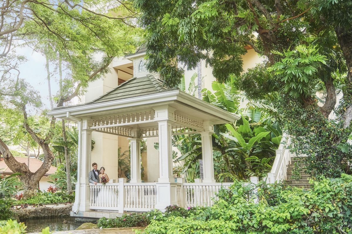 A man and woman in wedding attire stand in a white gazebo that overlooks a pond and is surrounded by greenery and next to a staircase.