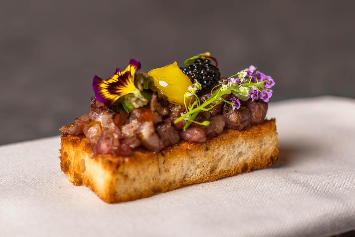 A square of toast topped with chopped Wagyu, caviar, a dollop of yellow sauce and edible flowers.