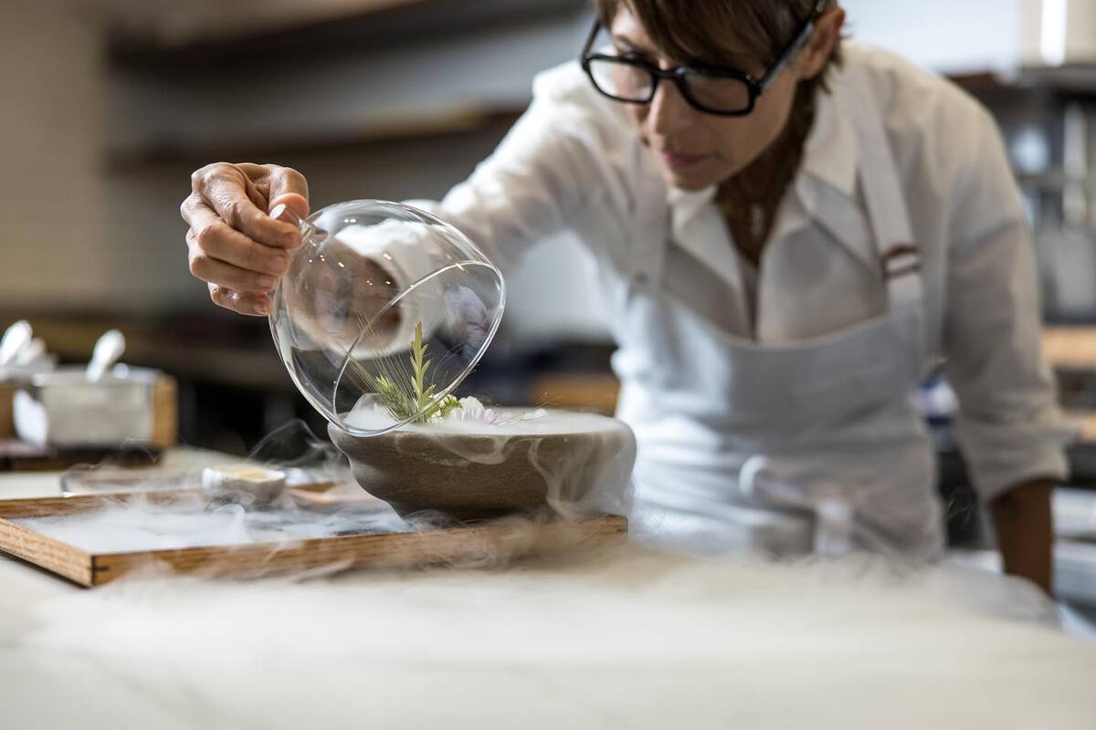 A female chef leans over and lifts a glass bell off a bowl that's smoking from dry ice.