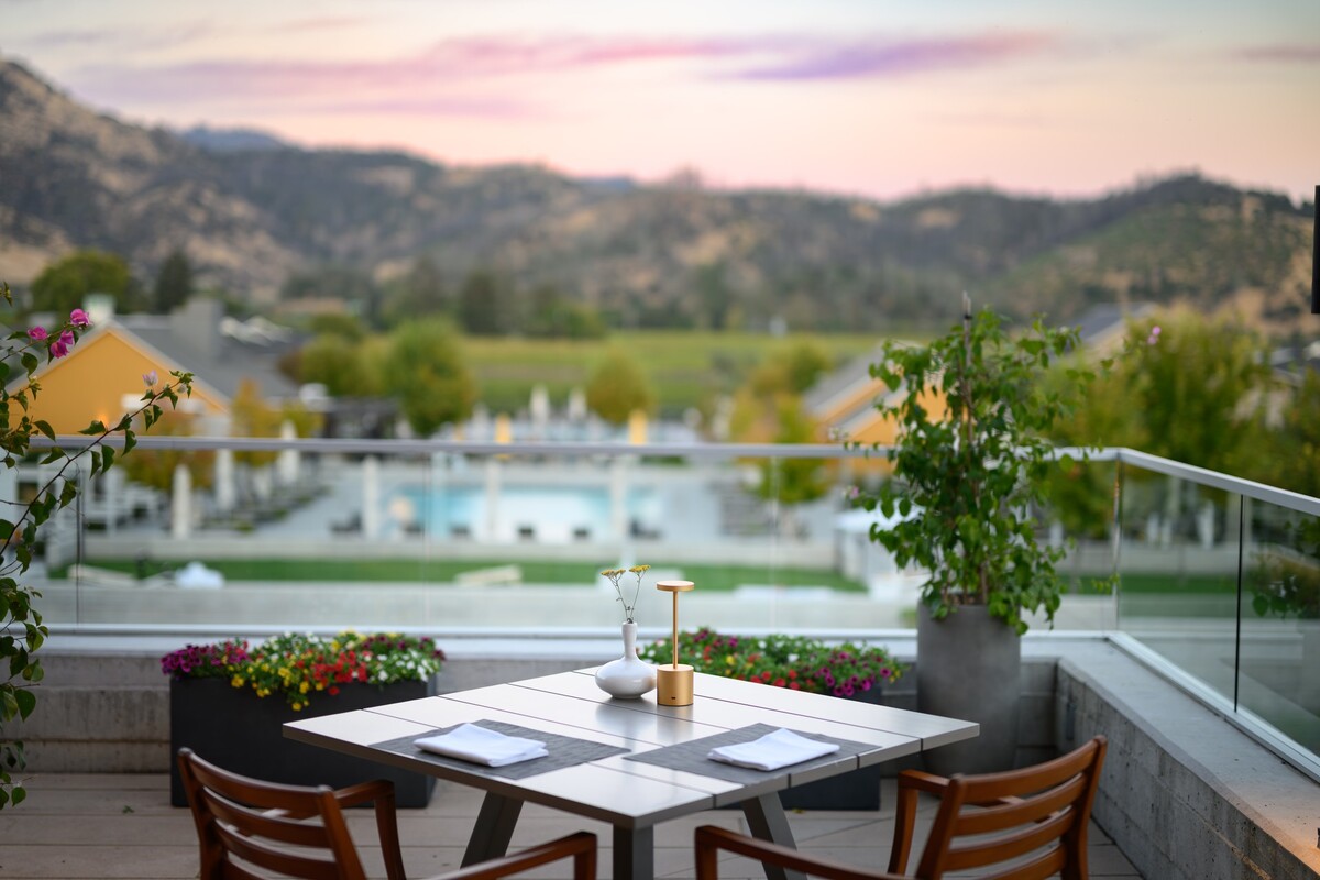 A set table sits on a balcony overlooking a sunset above a pool and hotel.