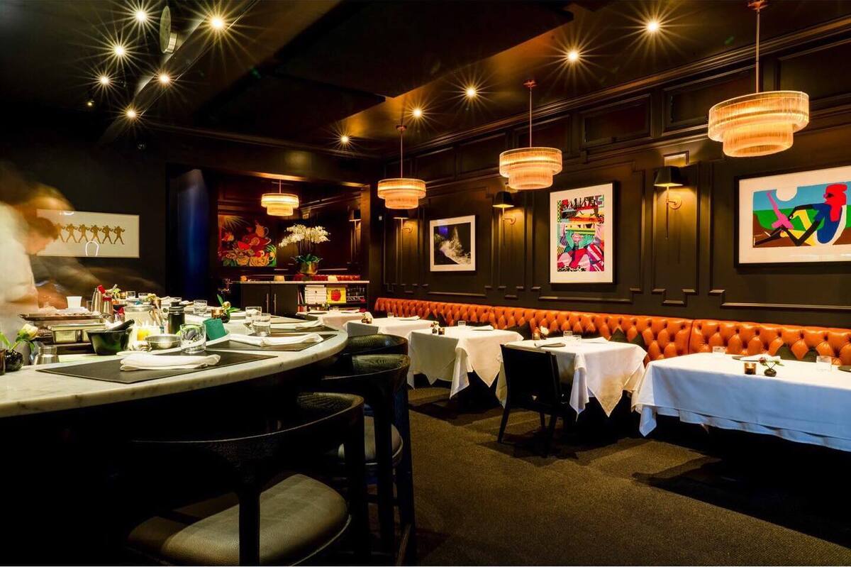 A dimly lit restaurant with a rounded bar, orange upholstered booths, hanging lights and colorful art against dark colored walls.