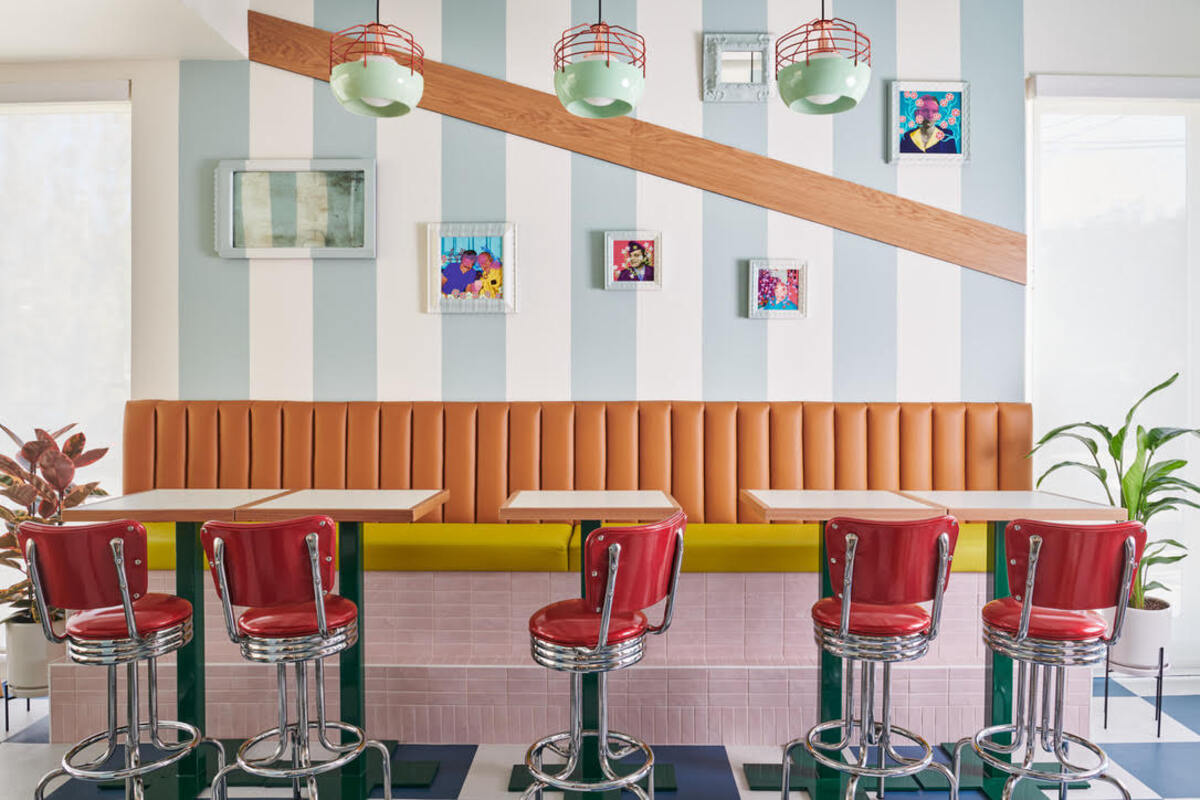 A restaurant with blue and white striped walls, orange and yellow booths, small tables and red and silver retro chairs.