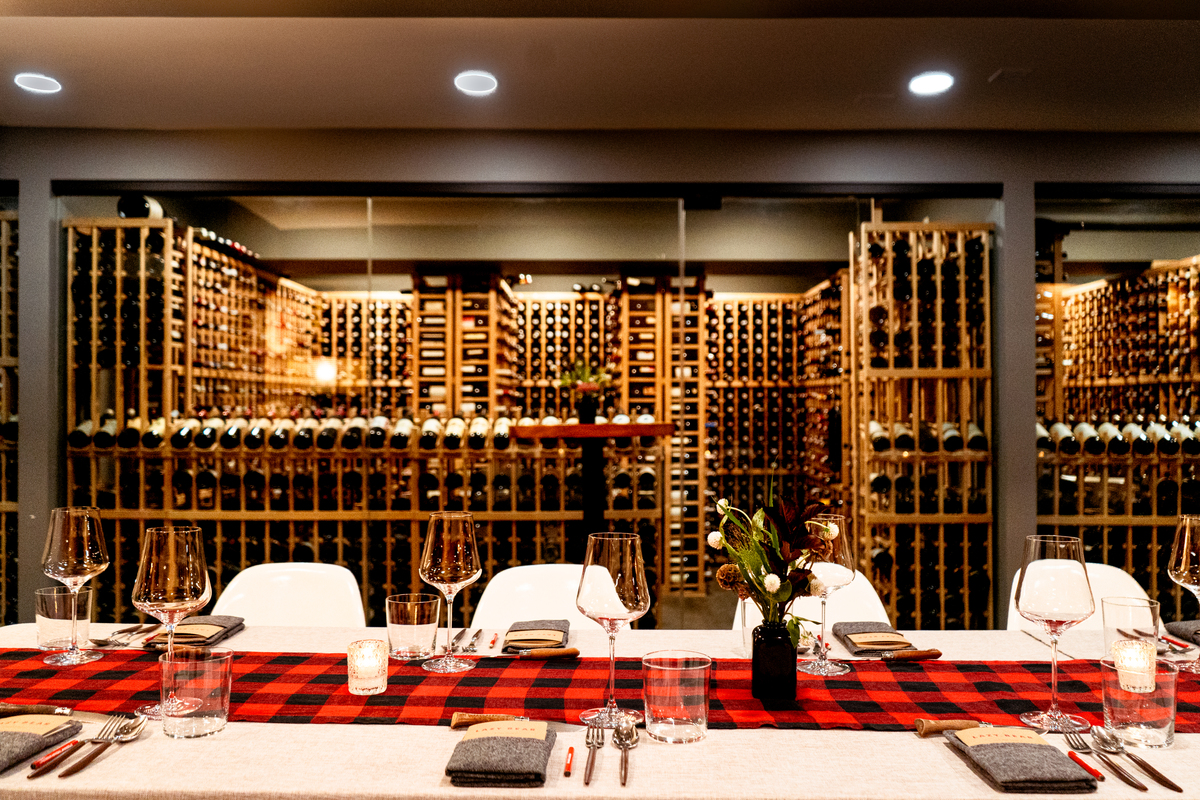 A long table with a black and red plaid table runner, wine glasses, utensils and menus against a backdrop of wine bottle storage.
