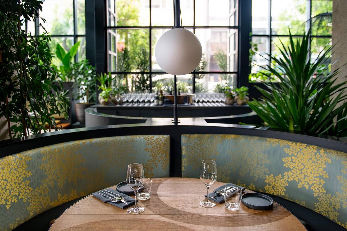 A round wooden table in a rounded both upholstered with green and blue flower fabric, a white bulb hanging light above and lots of surrounding plants.
