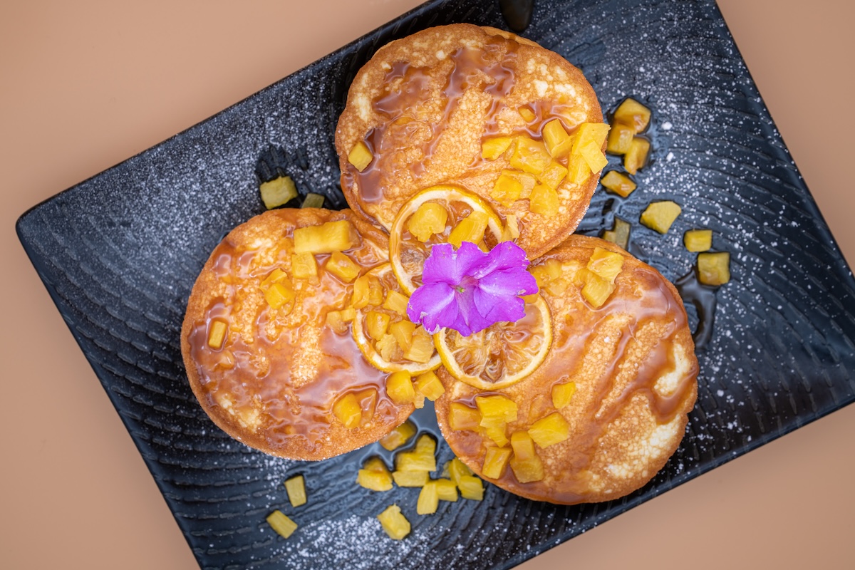 An overhead shot of three pancakes topped with chopped mango, syrup, orange slices and a purple flower.