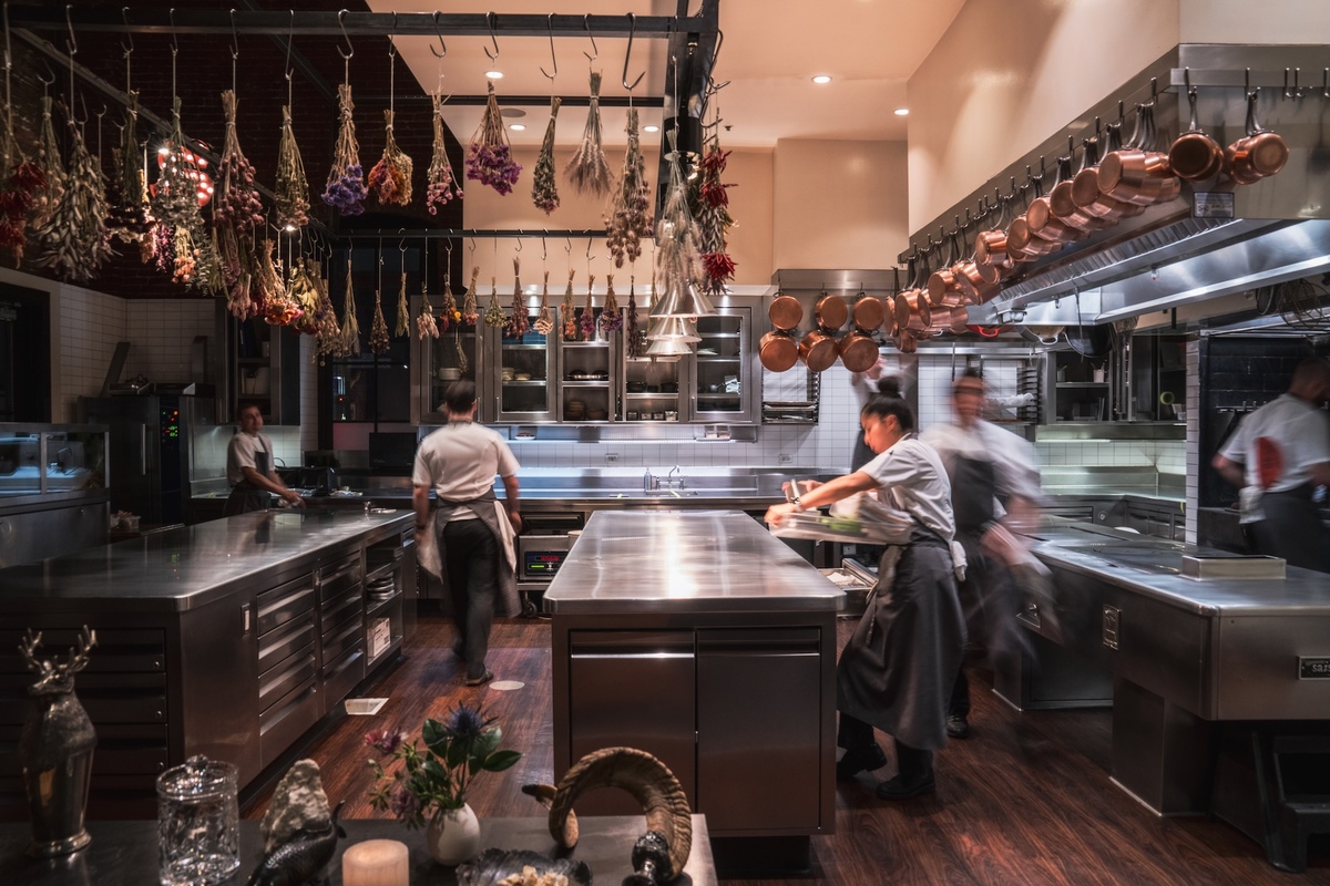 The view of a kitchen with stainless steel counters and appliances, hanging bouquets of dried flowers and copper pots while chefs rush about.
