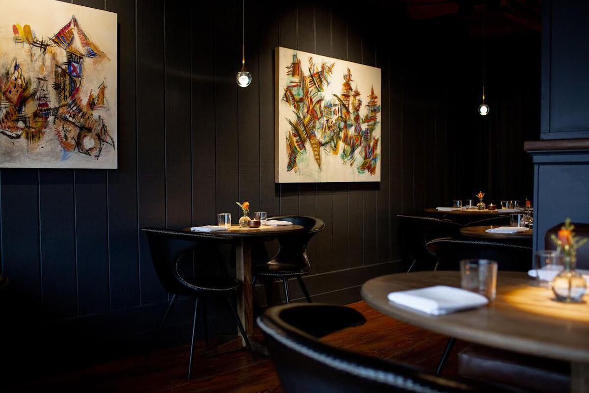 A dimly lit restaurant with black paneled walls, artwork and small tables lit by just an overhead light hanging above each.