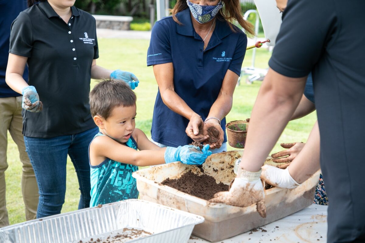 A kid and adults wearing a polo shirt uniform form balls of clay from tubs with it.