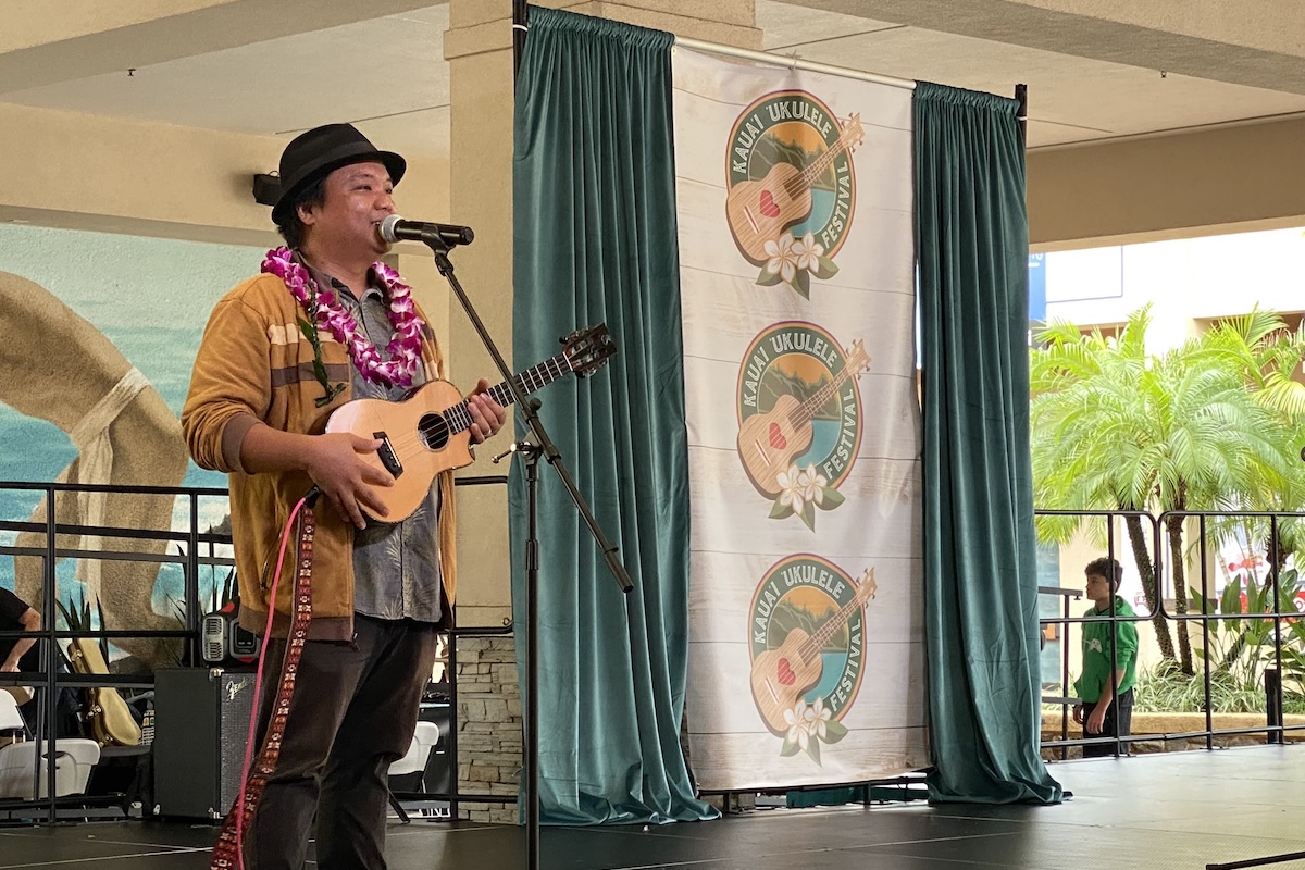 A man wearing a fedora and a lei plays a ukulele on stage.