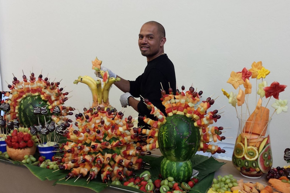 A man stands behind a table display of edible fruit arrangements.