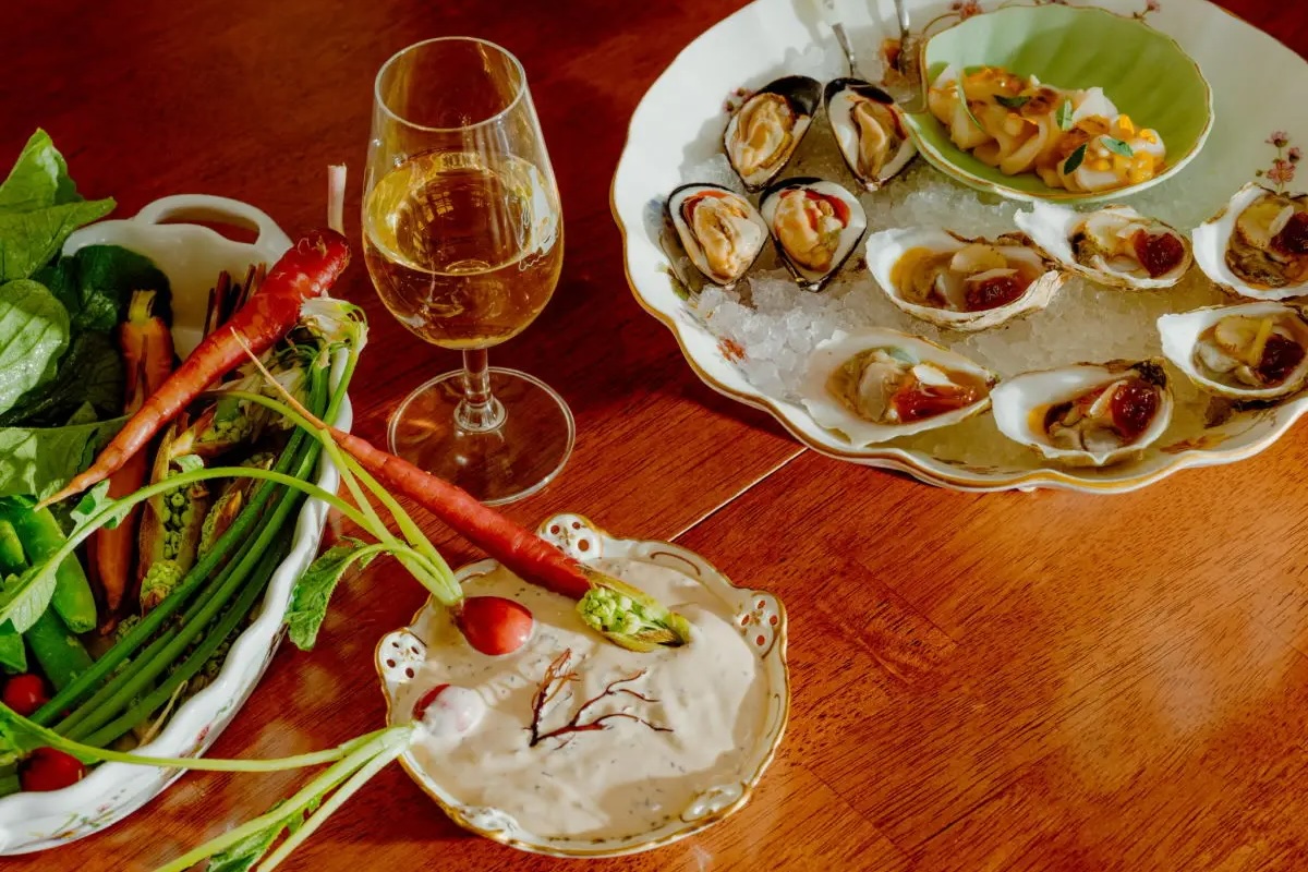 A wooden table displays a gourmet meal reminiscent of offerings at Oakland Michelin restaurants, featuring a glass of white wine, a bowl of assorted vegetables including carrots and radishes, a plate of oysters on ice with lemon wedges, and a small dish of dipping sauce.