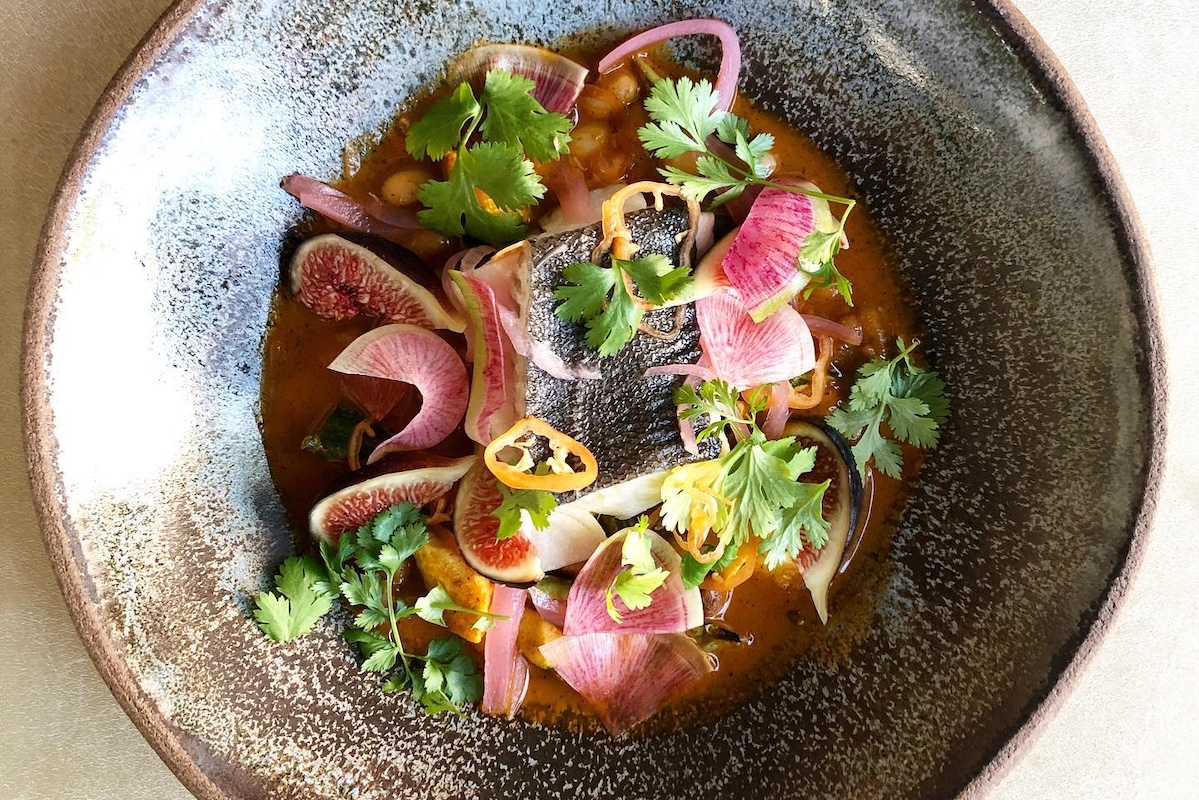 A bowl with soup topped with sliced figs and guava, cilantro, and fish.