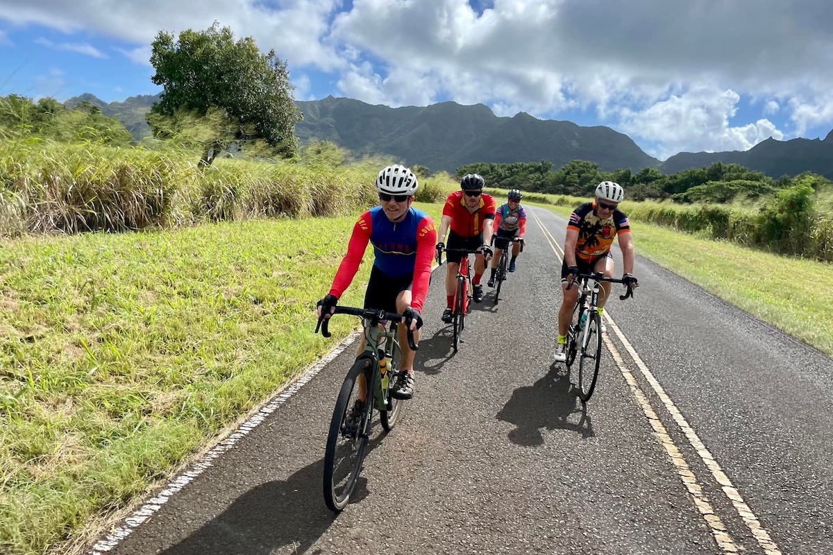 Four cyclists ride their bikes down a paved road with lots of green grass, hills and a cloudy sky for background.