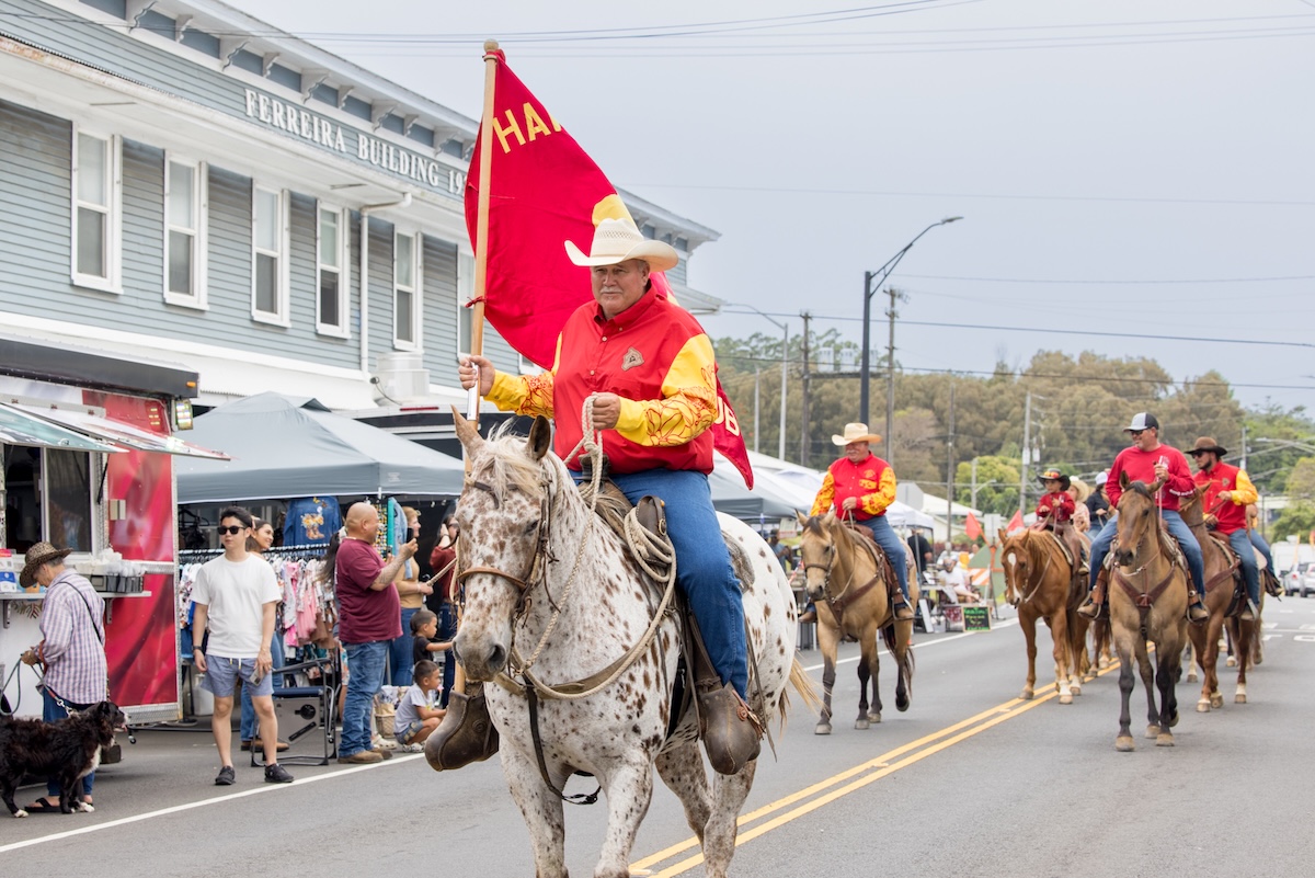 A man wearing a red and yellow long sleeve shirt and a cowboy hat holds a reg flag and rides a horse down the street as people watch on the sidewalks and others on horseback ride behind him.