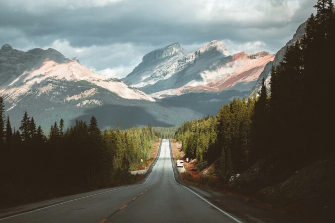 A photo of a road as it extends into the distance with gray mountains in the background, a cloudy sky and forest along the sides of the road.