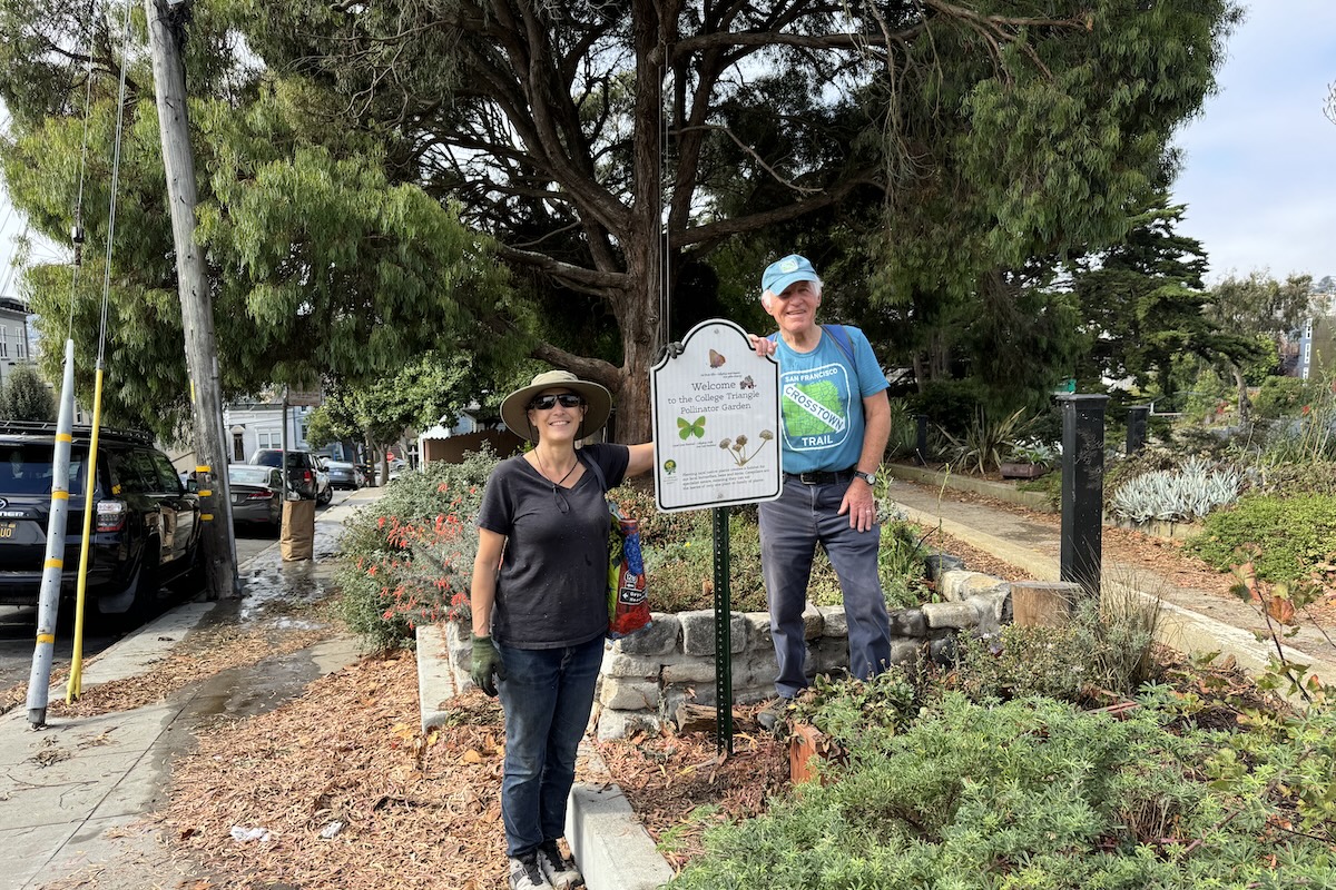 A woman and a man stand on either side of a sign amidst some greenery and bark.