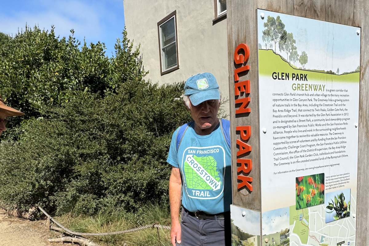 A hiker stands next to a trailhead sign with a map and red lettering that reads "Glen Park."