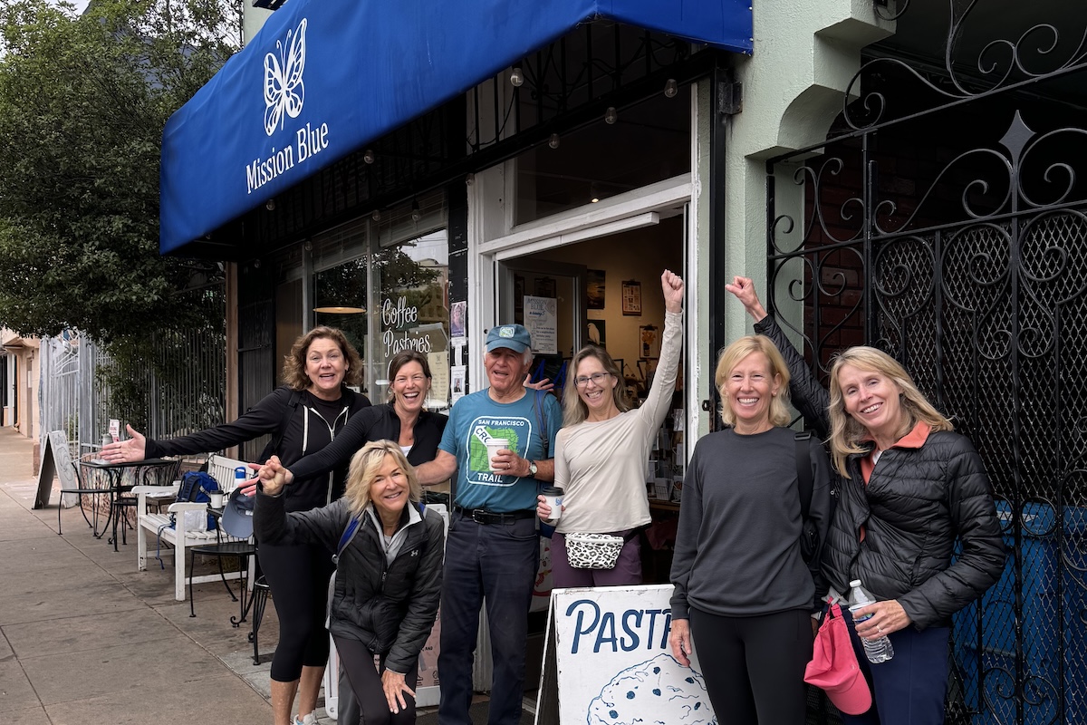 A group of hikers stands outside a coffee shop with a blue awning that reads "Mission Blue Coffee."