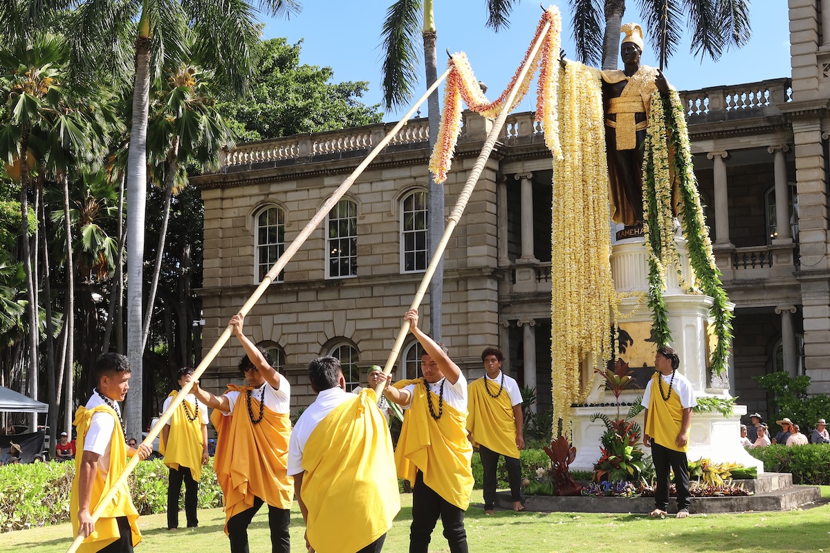 A group of men wearing white and yellow cultural attire use long poles to lift long leis onto the arms of a tall statue.