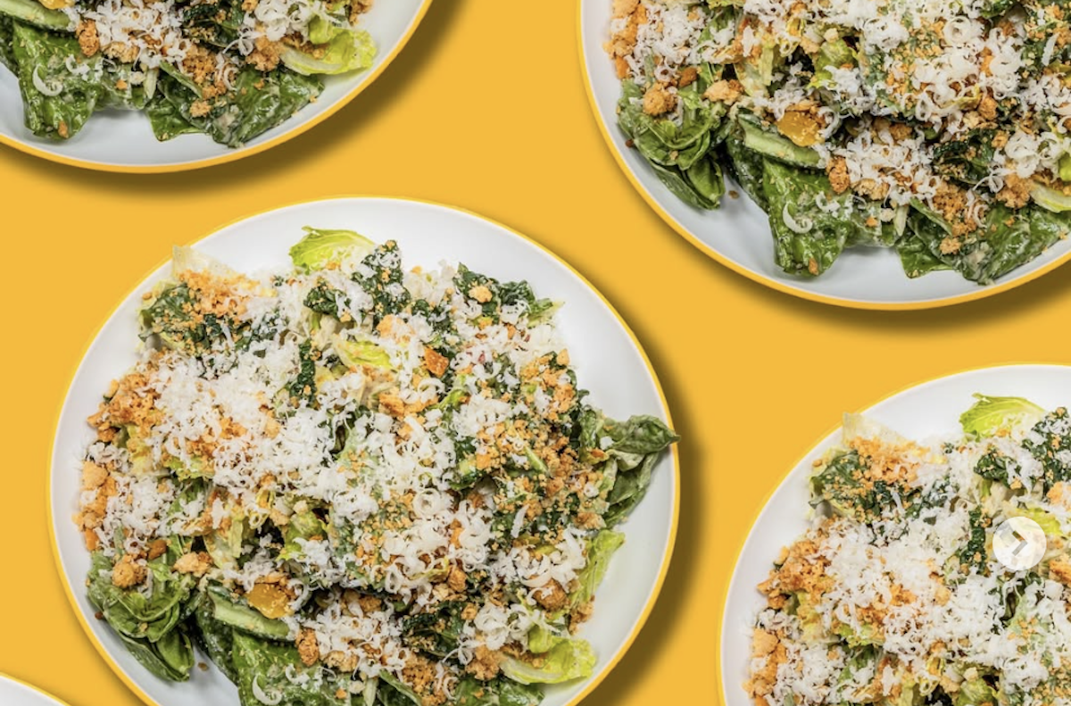 Plates of Caesar salad with grated cheese and breadcrumbs sits against a yellow background.