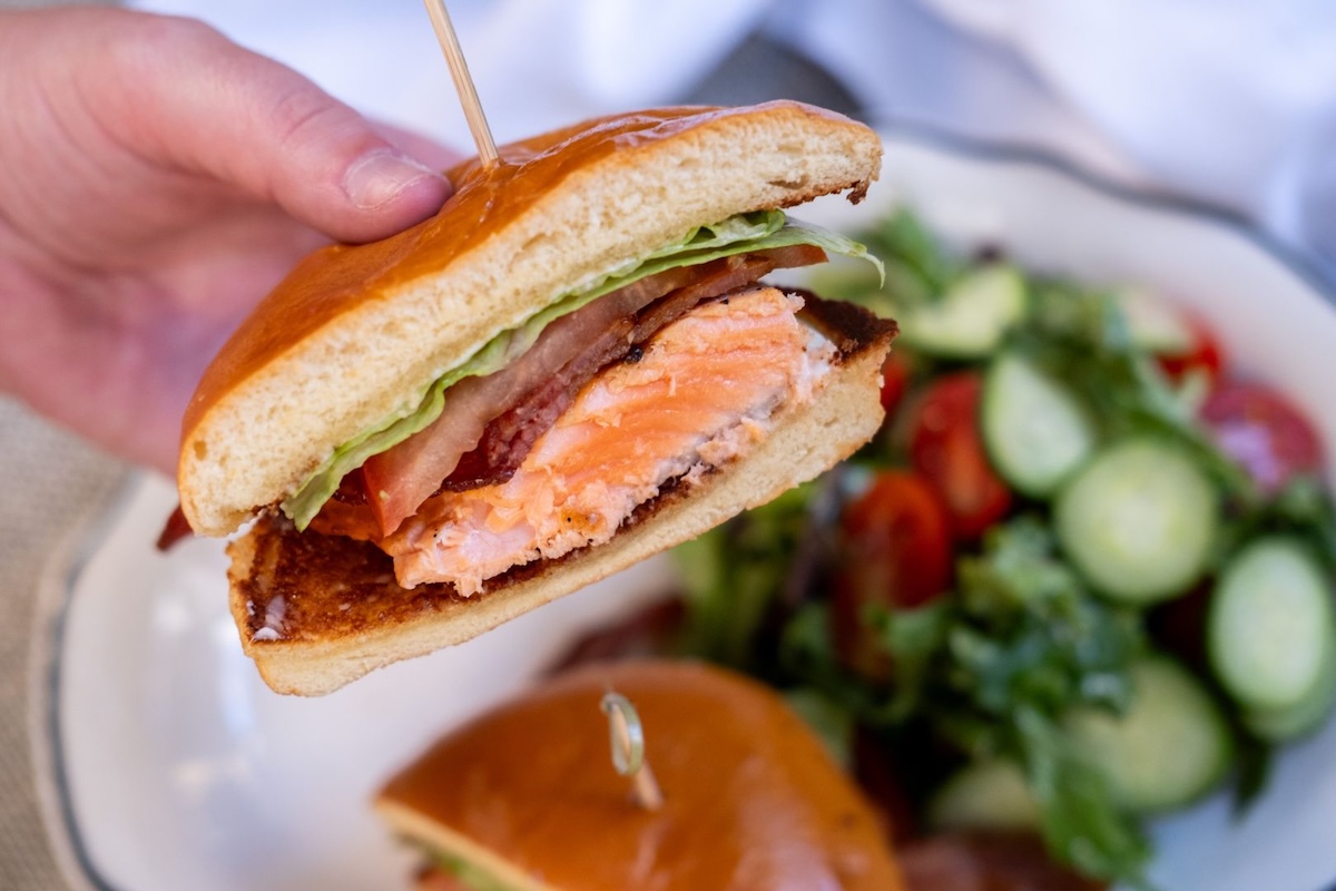 A person holds half of a salmon sandwich above a plate with the other half and a salad.