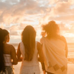 At sunrise, four women stand on the beach, creating a picturesque scene with the glowing horizon.