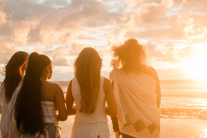 At sunrise, four women stand on the beach, creating a picturesque scene with the glowing horizon.