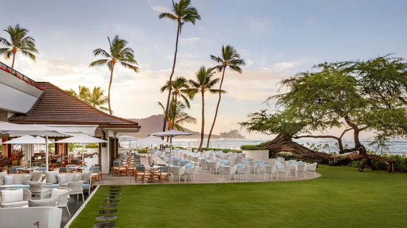 House-Without-a-Key-Oahu-Sunset-Dining-@halekulani-1600×800-1.png A scenic outdoor dining area with white tables and chairs on a grassy lawn, overlooking the ocean. Palm trees sway gently, and a distant mountain range is visible against a sunset sky. An adjacent building with a tiled roof is also part of the setting.