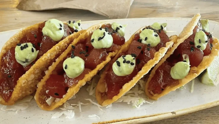 A plate from Best Poke Maui features four crispy taco shells filled with raw diced tuna topped with dollops of green avocado or wasabi cream and sprinkled with black sesame seeds. Shredded cabbage lies underneath the tacos, with a wedge of lime on the side.