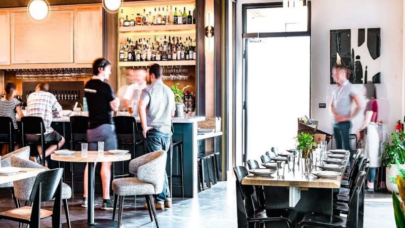 A modern restaurant offering the Best Dinner in Sacramento with a sleek, minimalist interior featuring wooden tables, cushioned chairs, and a well-lit bar area stocked with various bottles. Several people are engaging in conversations, creating a lively and welcoming atmosphere.