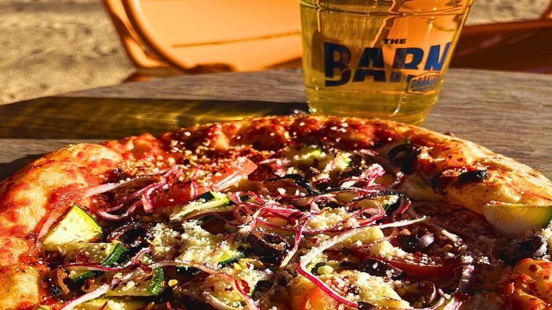 Close-up of a freshly baked pizza topped with vegetables, olives, red onions, and shredded cheese, placed on a wooden table. A glass of light beer with a frothy head sits next to the pizza, featuring a logo that reads "THE BARN," one of the best dining options near stadiums in Sacramento. Sunlit chairs are in the background.
