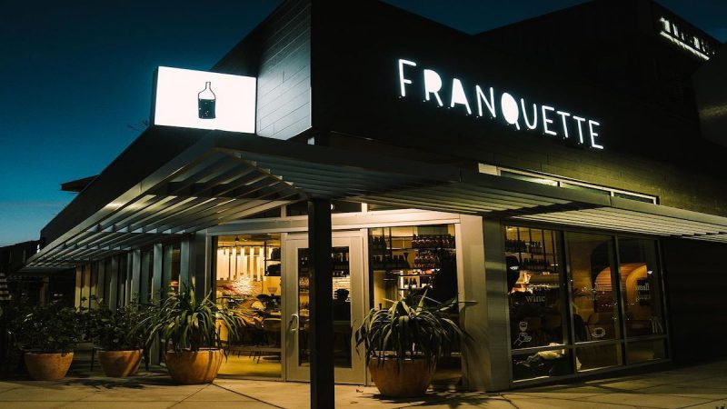 A modern restaurant named 'Franquette' is lit up at night. The exterior features large glass windows, offering a glimpse of the warm interior. Green plants in pots line the entrance, and a sign with a wine bottle icon glows above the doorway, making it one of the best dining spots near stadiums in Sacramento.