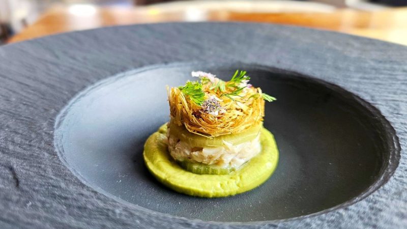 A small, elegantly plated dish, one of the best dinners in Sacramento, sits centered in a black bowl. It consists of a circular base of green puree, topped with a creamy mixture and garnished with a delicate, crispy nest-like structure and sprigs of herbs. The background is softly blurred.