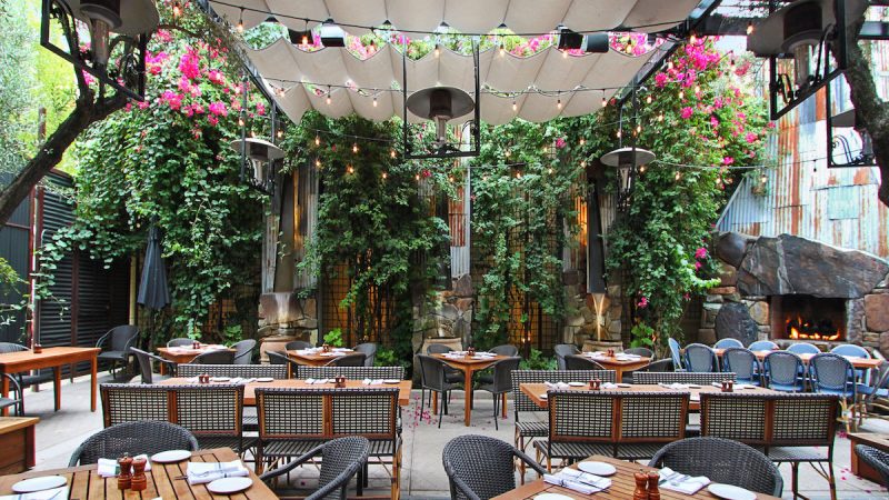 A charming outdoor dining area features rustic wooden tables and woven chairs, set under a canopy of fabric shades and surrounded by lush greenery and blooming pink flowers. Warm lighting and cozy decor create a welcoming and intimate atmosphere, making it the best dinner in Sacramento.