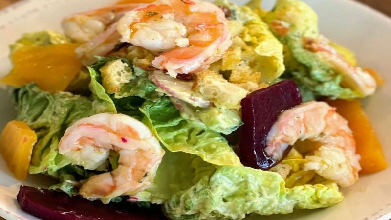 A plate of shrimp salad with mixed greens, sliced beets, and chunks of yellow fruit, likely mango or yellow beet. The salad is dressed with a creamy dressing and garnished with croutons. This delightful dish, often hailed as the best dinner in Sacramento, is served on a white plate placed on a wooden table.