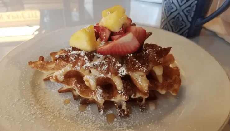 A plate with two stacked waffles topped with powdered sugar, fresh strawberries, and mango pieces. A patterned mug sits in the background, partially visible.