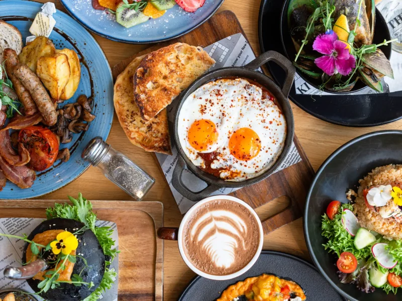 An overhead view of a table filled with various brunch dishes, including eggs and toast, a skillet with sunny-side-up eggs, a latte with latte art, a salad, and a bowl of mussels. The dishes are garnished with fresh herbs and edible flowers. Share your spread using #BrunchGiveaway for a chance to win!