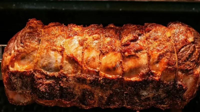Close-up of a large, perfectly roasted piece of meat on a rotisserie, with a golden-brown, crispy exterior that looks well-seasoned. The meat is secured with twine, showing an even, uniform cook with visible juices and caramelization.